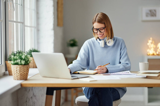 Щаслива молода жінка в окулярах переглядає дані на ноутбуці і робить нотатки в блокноті під час роботи над віддаленим проектом вдома
 - Фото, зображення