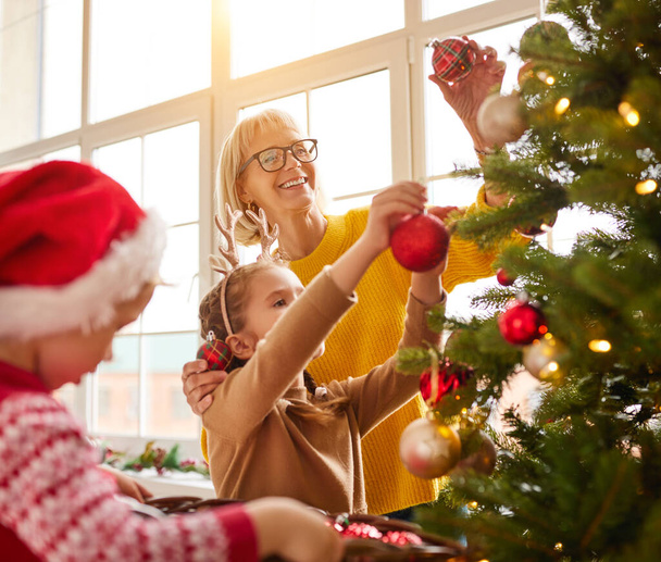 Merry mature woman and adorable children decorating coniferous Christmas tree with various baubles and ornament - 写真・画像