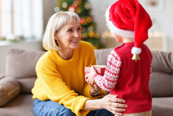 Fröhliche Großmutter bekommt Weihnachtsgeschenk von bis zur Unkenntlichkeit ergrautem kleinen Enkelkind, während sie zu Hause Urlaub feiert - Foto, Bild