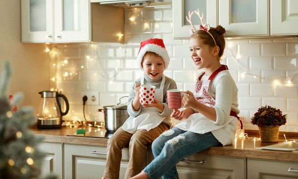 szczęśliwy śmieszne dzieci w Wigilię, dziewczyna i chłopiec śmiać się i pić gorący napój kakaowy, które upiekli razem w przytulnej kuchni w hom - Zdjęcie, obraz