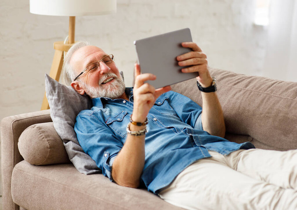 Optimistic bearded elderly male relaxing on sofa and browsing tablet on weekend day at hom - 写真・画像