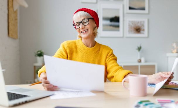 スタイリッシュなカジュアルな服を着た陽気な高齢者の女性は笑顔とテーブルに座って家で働く中で文書を調べる - 写真・画像