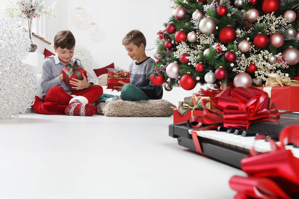 merry christmas and happy holidays, children look at the gift packages near the christmas tree with musical instruments, at home in the living room sitting on the floor - Фото, изображение
