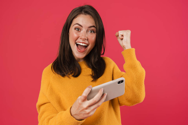 Image of excited girl making winner gesture while playing video game on cellphone isolated over pink background - Фото, изображение
