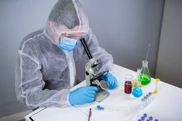 Φαρμακευτικοί παρασκευαστές αναπτύσσουν ιογενή φάρμακα εφευρίσκοντας το καλύτερο συστατικό. Επιστήμονας που φοράει ατομικό προστατευτικό εξοπλισμό με μάσκα προσώπου που παρακολουθεί τα αποτελέσματα κυττάρων στο μικροσκόπιο στο εργαστήριο - Φωτογραφία, εικόνα