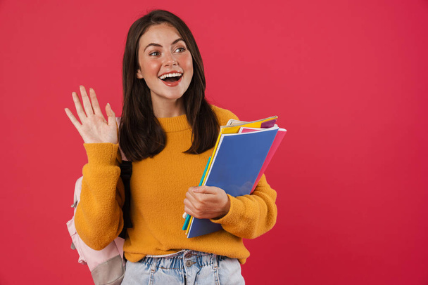 Image of happy student girl waving hand while holding exercise books isolated over pink background - Φωτογραφία, εικόνα