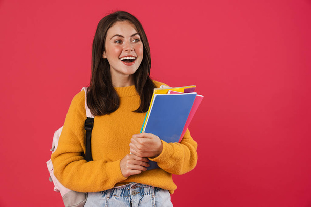 Χαρούμενη νεαρή μαθήτρια που φοράει σακίδιο, κουβαλάει βιβλία που στέκονται απομονωμένα σε ροζ φόντο - Φωτογραφία, εικόνα