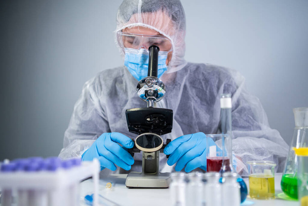 Φαρμακευτικοί παρασκευαστές αναπτύσσουν ιογενή φάρμακα εφευρίσκοντας το καλύτερο συστατικό. Επιστήμονας που φοράει ατομικό προστατευτικό εξοπλισμό με μάσκα προσώπου που παρακολουθεί τα αποτελέσματα κυττάρων στο μικροσκόπιο στο εργαστήριο - Φωτογραφία, εικόνα
