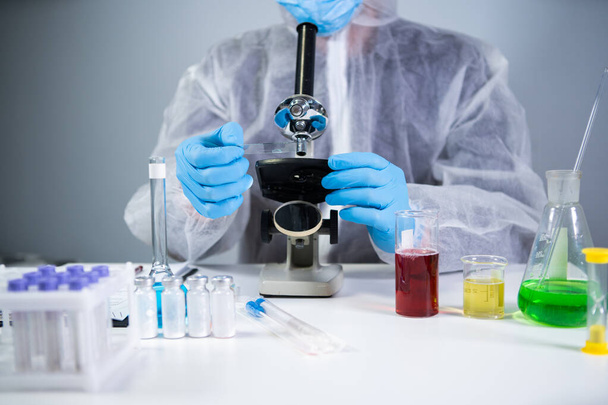 Bilim adamı mikroskopla numune inceliyor, bilim laboratuarında tıbbi araştırma yapıyor. Koruyucu giysiler içindeki erkek laboratuvar asistanı laboratuvarda mikroskopla yeni kimyasal maddeler üzerinde çalışıyor.. - Fotoğraf, Görsel