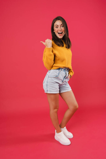 Full length portrait of a happy casual woman pointing finger away isolated on pink background - Foto, Imagem