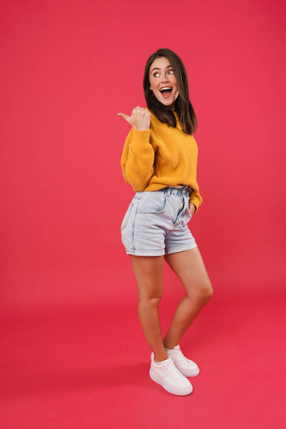 Full length portrait of a happy casual woman pointing finger away isolated on pink background - Foto, imagen