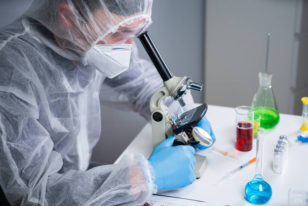 顕微鏡でサンプルを調べる科学者、科学研究所で医学研究を行う。研究室で顕微鏡を通して新しい化学物質を研究する保護服の男性研究室のアシスタント. - 写真・画像