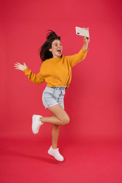 Image of excited brunette girl taking selfie photo on cellphone while jumping isolated over pink background - Valokuva, kuva