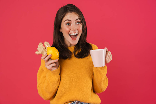 Image of excited caucasian girl holding lemon and ginger while drinking tea isolated over pink background - Valokuva, kuva