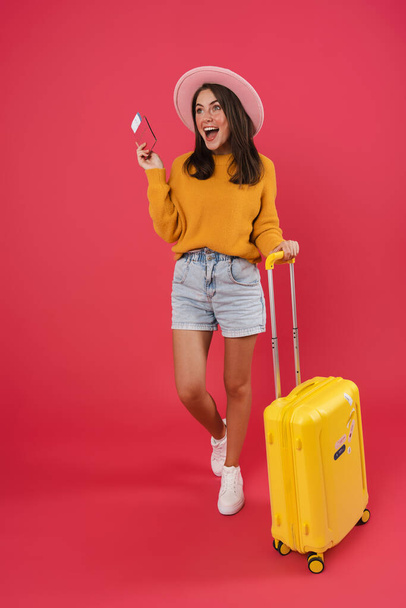 Full length of a happy young female walking with the travel bag, holding passport and tickets isolated over pink background - Fotografie, Obrázek