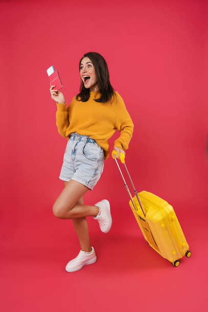 Full length of a happy young female walking with the travel bag, holding passport and tickets isolated over pink background - Foto, Imagem