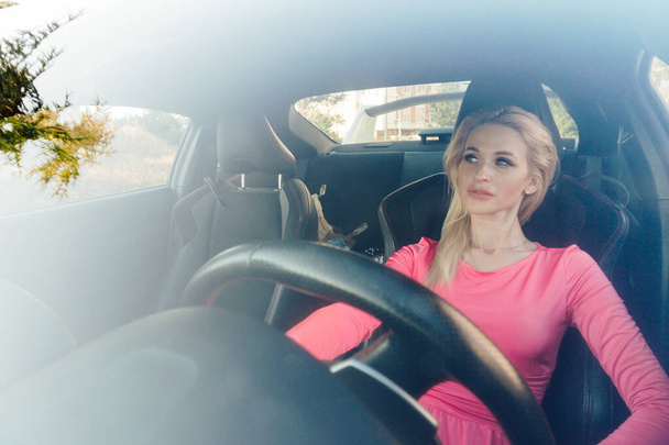 Όμορφη ξανθιά γυναίκα σε ροζ φόρεμα πίσω από το τιμόνι ενός αυτοκινήτου - Φωτογραφία, εικόνα
