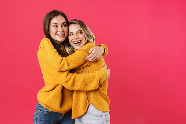 Portrait of two young female friends standing over pink background, hugging - Foto, Imagem