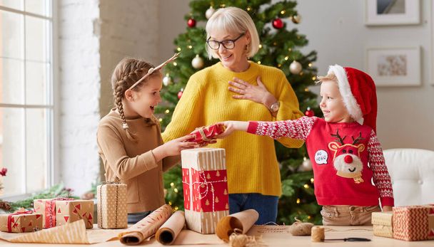 счастливая семья: улыбающаяся бабушка и смешные внуки смеются и вместе обернутые подарок, стоя в уютной комнате с Рождеством тр - Фото, изображение