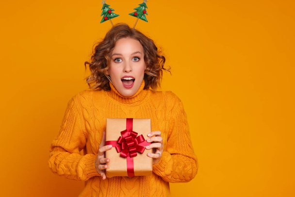 alegre divertida sorprendida joven mujer en un suéter amarillo con la boca abierta sostiene un regalo de Navidad en un fondo amarillo de color - Foto, Imagen