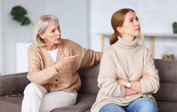 Serious aged mother touching chin and looking at depressed adult daughter while sitting on couch after argument at hom - Фото, изображение