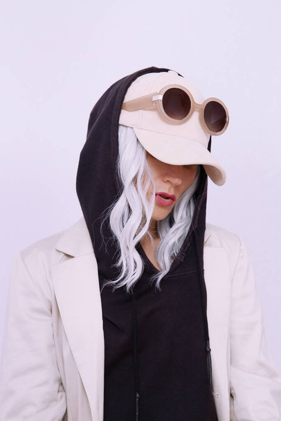 Blond meisje in een witte studio. Trendy beige casual outfit met zwarte hoodie details. Cap, zonnebril, spijkerjasje en broek. Straatstijl. Herfst winterseizoen. Modelookbook concept - Foto, afbeelding