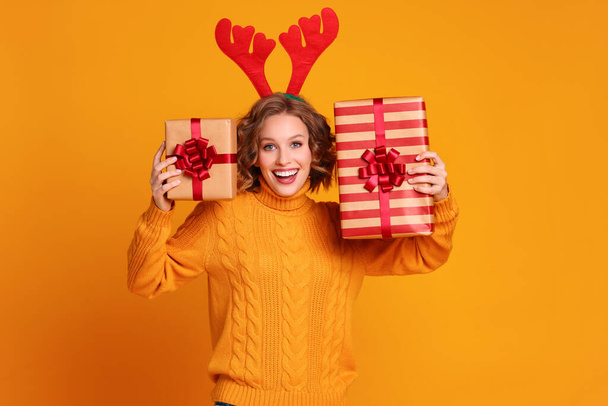 χαρούμενη αστεία έκπληξη νεαρή γυναίκα σε ένα κίτρινο πουλόβερ με κόκκινα κέρατα ελαφιού κατέχει ένα χριστουγεννιάτικο δώρο και γέλιο σε ένα χρωματιστό κίτρινο backgroun - Φωτογραφία, εικόνα