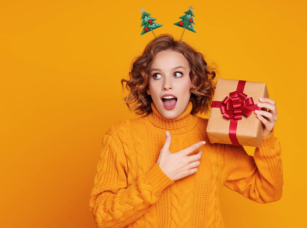 vidám vicces meglepett fiatal nő egy sárga pulóverben nyitott szájjal tart karácsonyi ajándékot egy színes sárga backgroun - Fotó, kép