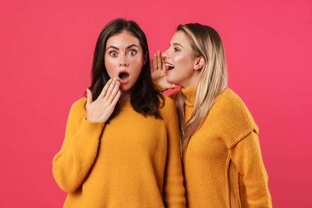 Image of beautiful woman whispering secret to shocked her friend isolated over pink background - Фото, изображение