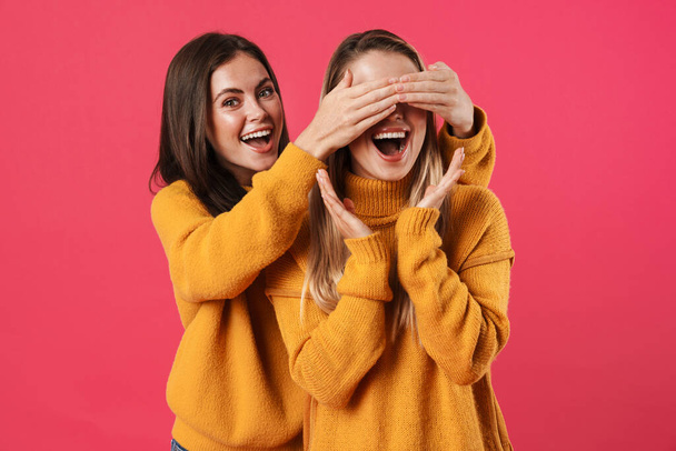 Image of happy beautiful woman covering eyes to her surprised friend isolated over pink background - Фото, зображення