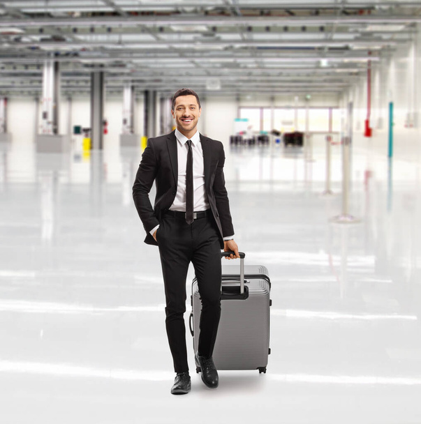 Full length portrait of a smiling young man in a black suit walking in an empty airport hall with a suitcase - Foto, Imagen