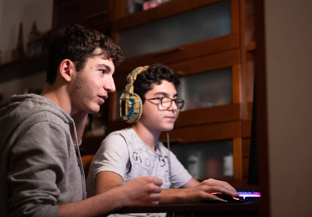 Rester à la maison : Deux frères caucasiens restent occupés avec leur jeu vidéo préféré. L'un est assis avec le casque de jeu sur sa tête et l'autre est debout à côté de lui pour suggérer des mouvements à prendre. - Photo, image