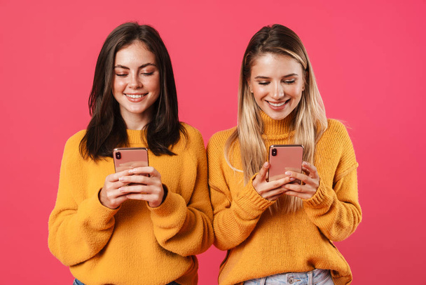 Image of joyful nice women smiling while using smartphones isolated over pink background - Foto, imagen