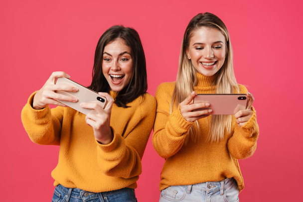 Image of delighted nice women playing video game on smartphones isolated over pink background - Photo, image