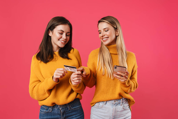 Two happy young girlfriends shopping online with mobile phone and credit card isolated on pink wall background - Valokuva, kuva