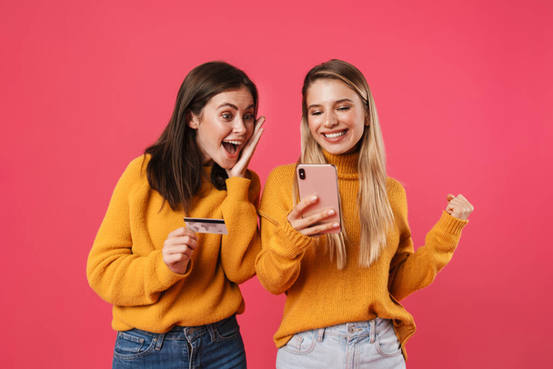 Two happy young girlfriends shopping online with mobile phone and credit card isolated on pink wall background - Zdjęcie, obraz