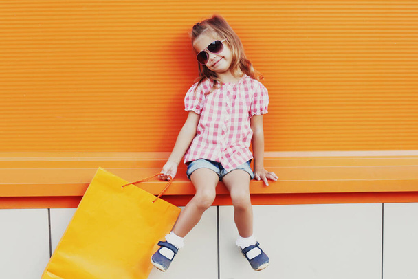 κοριτσάκι παιδί με τσάντες ψώνια στο δρόμο της πόλης πάνω από πορτοκαλί φόντο - Φωτογραφία, εικόνα