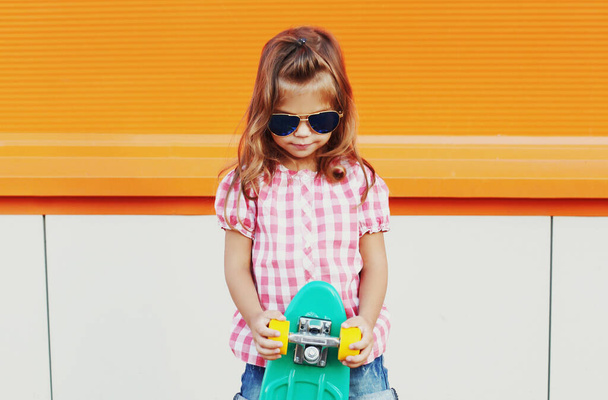 Portrait of stylish little girl child with skateboard in the city over an orange background - Photo, Image