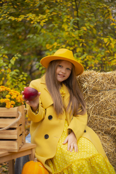 秋の飾りの横にあるオレンジの帽子、ドレス、コートのかわいい十代のブルネットの女の子は赤いリンゴを持っています。美人. - 写真・画像