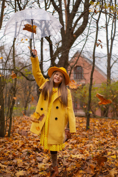 Nettes hübsches brünettes Mädchen im Teenageralter mit orangefarbenem Hut, Kleid und Mantel, das einen transparenten Regenschirm mit fallenden Blättern über sich hält. Gemütlichkeit, Herbst. - Foto, Bild