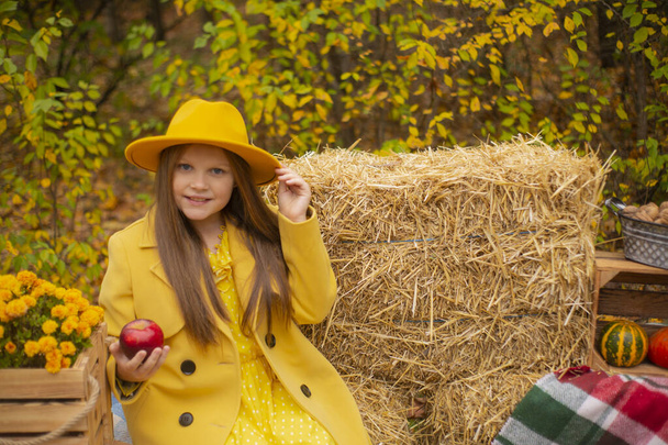 χαριτωμένο όμορφη έφηβη μελαχρινή κοπέλα σε ένα πορτοκαλί καπέλο, φόρεμα και παλτό δίπλα στο φθινόπωρο διακοσμήσεις κρατώντας ένα κόκκινο μήλο. Ανάγκη. - Φωτογραφία, εικόνα