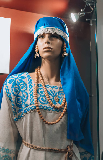 Traditionelle ukrainische Nationalkleidung im Khotyn Museum. Nahaufnahme Porträt der ukrainischen Schaufensterpuppe mit farbenfroher blauer Kopfbedeckung auf Kopf und Halskette. weißes Baumwollkleid - Foto, Bild