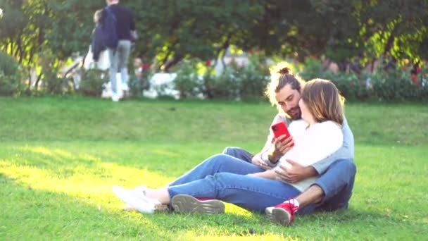 Onnellinen pari käyttää matkapuhelinta istuessaan nurmikolla, nainen nojaa mieheensä. Konsepti. Puhelinriippuvuus, mies surffaa netissä treffeillä tyttöystävänsä kanssa, - Materiaali, video
