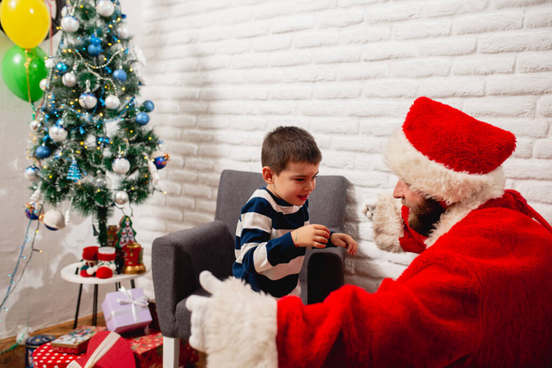Ένας νεαρός ντυμένος Άγιος Βασίλης με απλωμένα χέρια και ένα χαριτωμένο αγόρι τρέχει στην αγκαλιά του στο σπίτι δίπλα σε ένα στολισμένο χριστουγεννιάτικο δέντρο. Καλές γιορτές. - Φωτογραφία, εικόνα