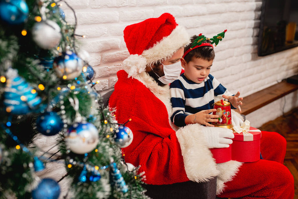 Seitenansicht eines süßen 5-jährigen Jungen, der mit Gesichtsmaske auf dem Schoß des Weihnachtsmannes sitzt und neben einem geschmückten Weihnachtsbaum ein Weihnachtsgeschenk betrachtet. Winterurlaub während der Pandemie COVID -19 Coronavirus - Foto, Bild