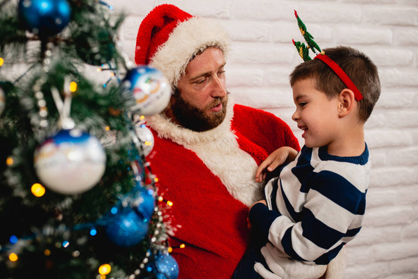 部屋の装飾された青いクリスマスツリーの横にある笑顔ひげを生やしたサンタクロースの腕の中で彼の頭の上に小道具を持つ美しい小さな男の子。お正月とクリスマス - 写真・画像