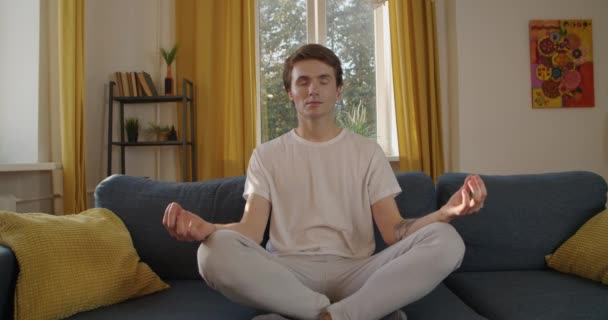 Jongeman zittend op de bank yoga ontspanning oefenen na drukke werkweek slow motion. Vooraanzicht van aantrekkelijke persoon mediteren alleen thuis achtergrond. Anti-stress oefening spirituele levensstijl - Video