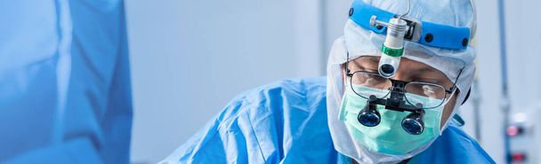 Aziatische arts Chirurg in masker dragen chirurgische loupes tijdens de medische procedure, Chirurgie operatiekamer met elektrocauterisatie apparatuur voor cardiovasculaire spoedoperatie centrum, banner achtergrond. - Foto, afbeelding
