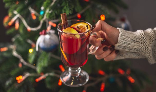 A woman's hand in a warm sweater is holding a cup of aromatic hot mulled wine against the background of a Christmas tree with lights. Concept of a festive atmosphere. - Foto, afbeelding