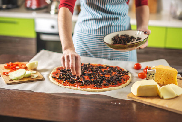 A female chef in an apron is putting mushrooms on a pizza raw. Cooking delicious pizza at home in the kitchen. - Photo, Image